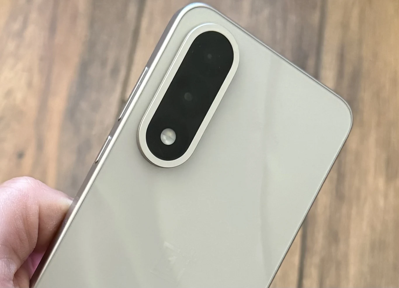 OnePlus Nord 5 ma podwójny aparat fotograficzny. Nowoczesny smartfon o jasnej obudowie z widocznym potrójnym aparatem umieszczonym w pionowej ramce, trzymany w dłoni na tle drewnianego blatu.