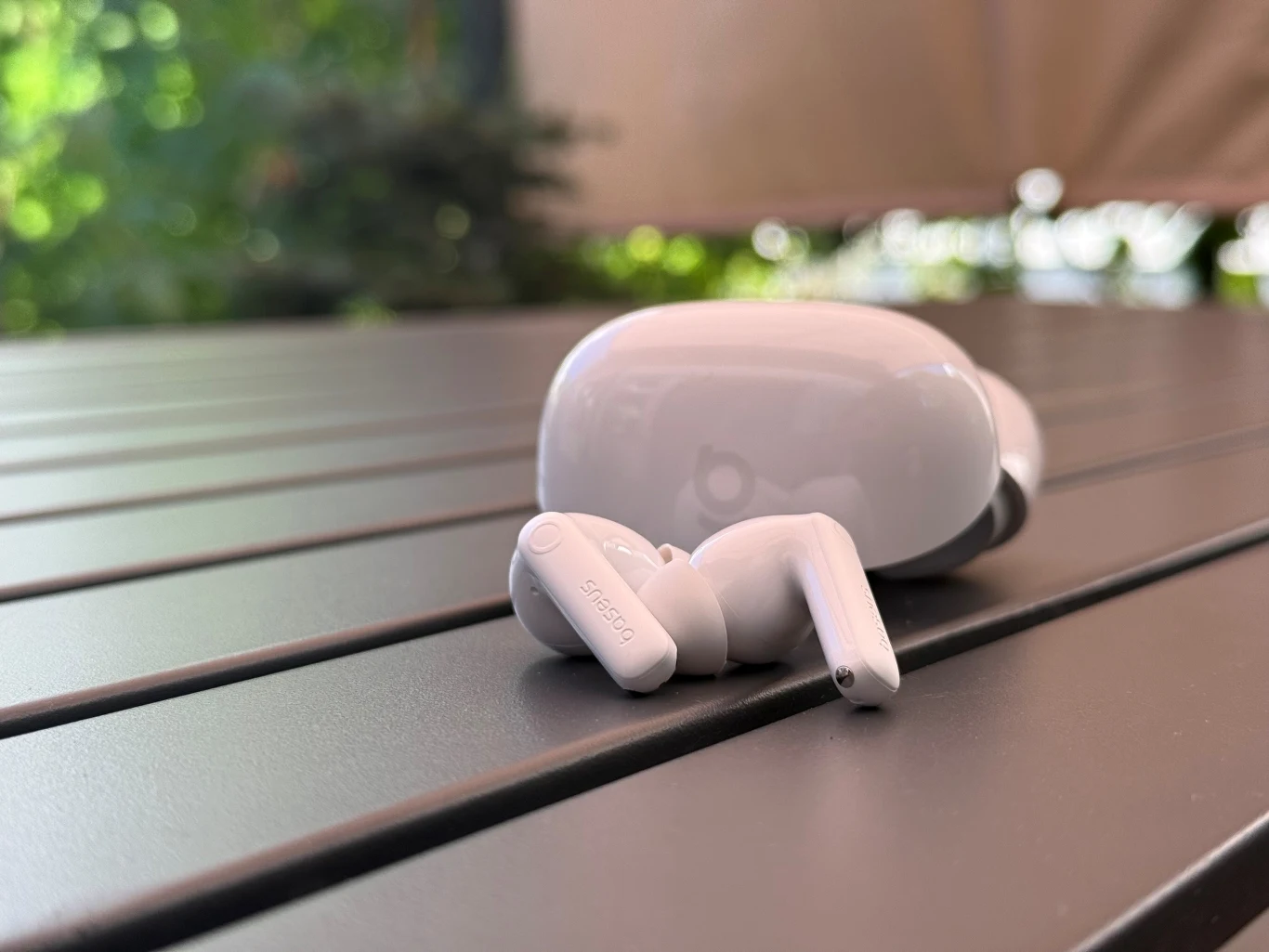 Baseus Bass BP1 Pro - budżetowe słuchawki, które zaskakują jakością Białe bezprzewodowe słuchawki douszne oraz etui ładujące leżące na drewnianym stole na tle rozmazanej zieleni i beżowej tkaniny.