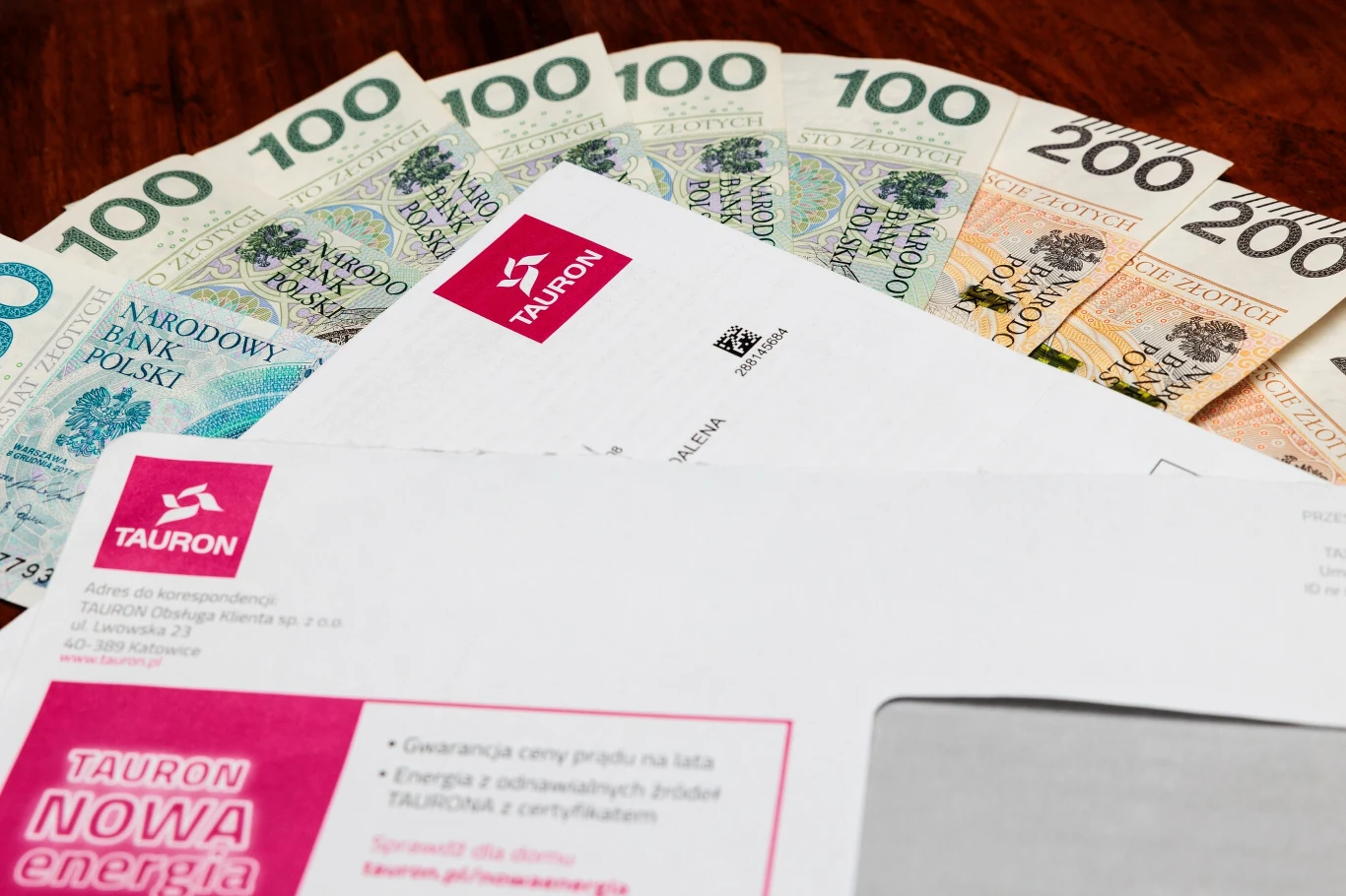 Rachunki bardziej przejrzyste Banknoty polskich złotych o nominałach 100 i 200 oraz koperta i rachunek firmy Tauron ułożone na drewnianym stole, symbolizujące opłacanie rachunków oraz kwestie finansowe związane z energią elektryczną.