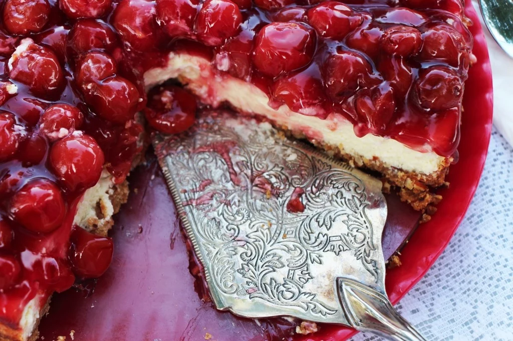 To ciasto nie jest skomplikowane, a z pewnością oczaruje i zachwyci wszystkich gości Sernik z grubą warstwą wiśni w żelu, podany na czerwonym talerzu z ozdobną łopatką do ciasta, z wyciętym kawałkiem.