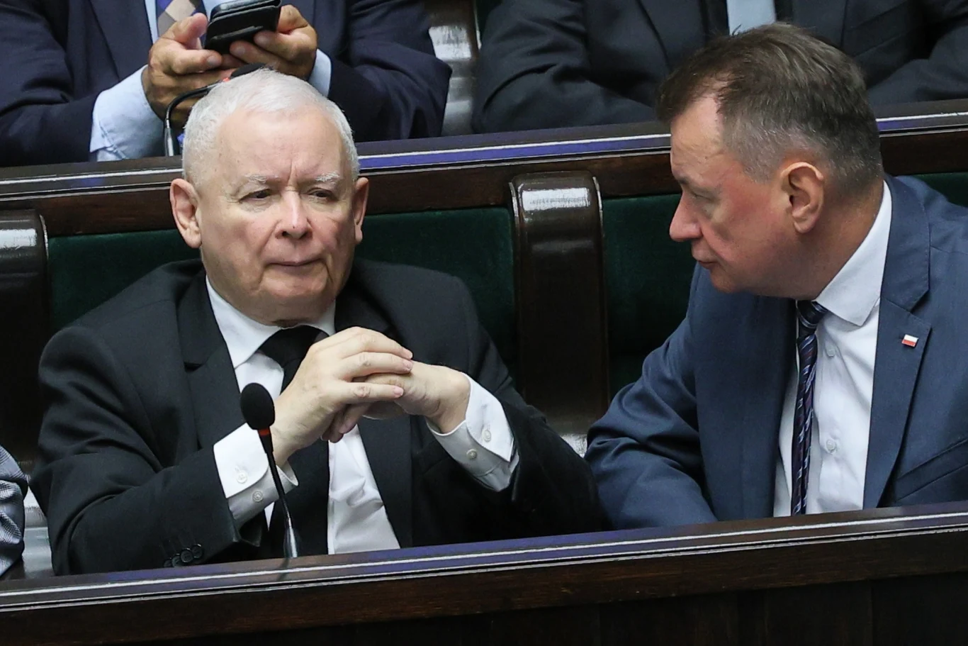 Prezes PiS Jarosław Kaczyński reaguje ws. sytuacji na granicy Dwóch mężczyzn w garniturach rozmawia ze sobą w ławach parlamentu, jeden z nich ma złożone ręce, drugi pochyla się w jego stronę, w tle widoczni inni uczestnicy obrad.
