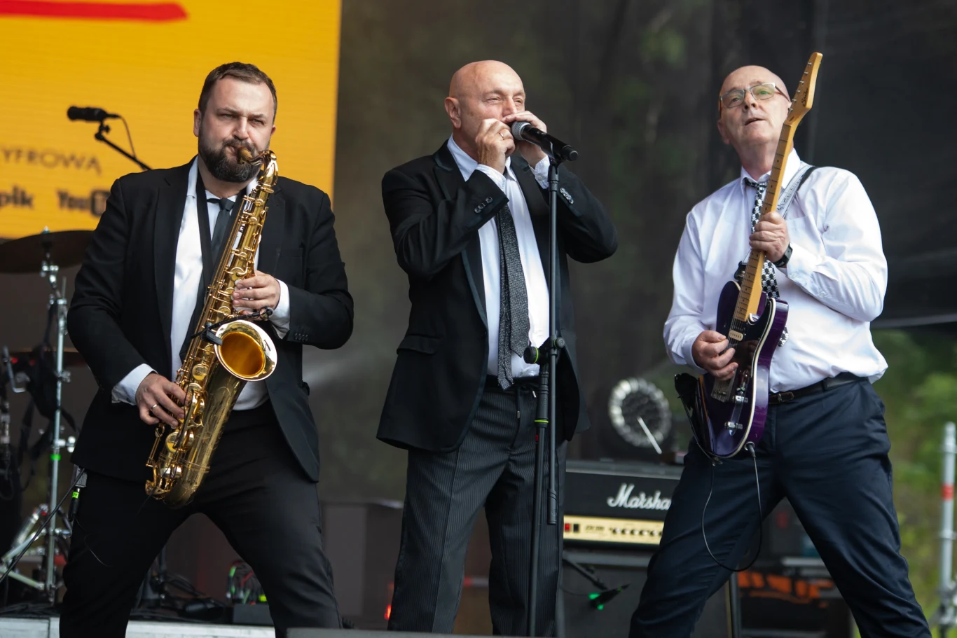 Trzech mężczyzn w garniturach występuje na scenie, jeden gra na saksofonie, drugi śpiewa do mikrofonu, trzeci gra na gitarze elektrycznej, w tle sprzęt muzyczny i kolorowa plansza na ekranie.