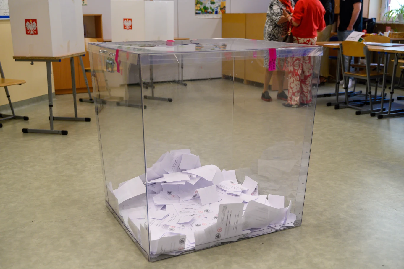Ponowne przeliczenie głosów. Polacy zabrali głos Duża, przezroczysta urna wyborcza wypełniona kartami do głosowania ustawiona na środku sali, w tle widoczne stanowiska do głosowania oraz kilka osób.