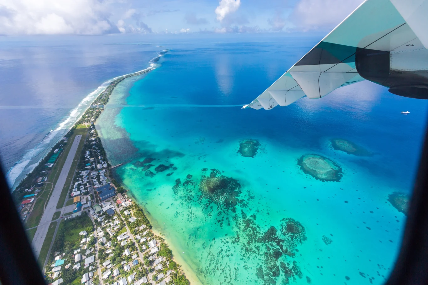 Malowniczy archipelag Tuvalu może znaleźć się pod wodą Widok z okna samolotu na wyspę z pasem startowym, zabudowaniami oraz turkusową lagunę otoczoną rafą koralową i przejrzystą wodą Oceanu Spokojnego, z fragmentem skrzydła samolotu w prawym górnym rogu.