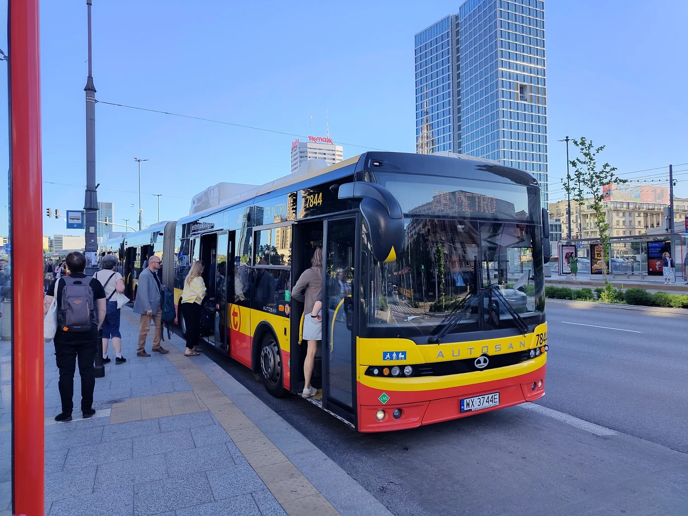 Miejski autobus z otwartymi drzwiami, do którego wsiadają i z którego wysiadają pasażerowie, na tle nowoczesnych wieżowców i centrum dużego miasta.