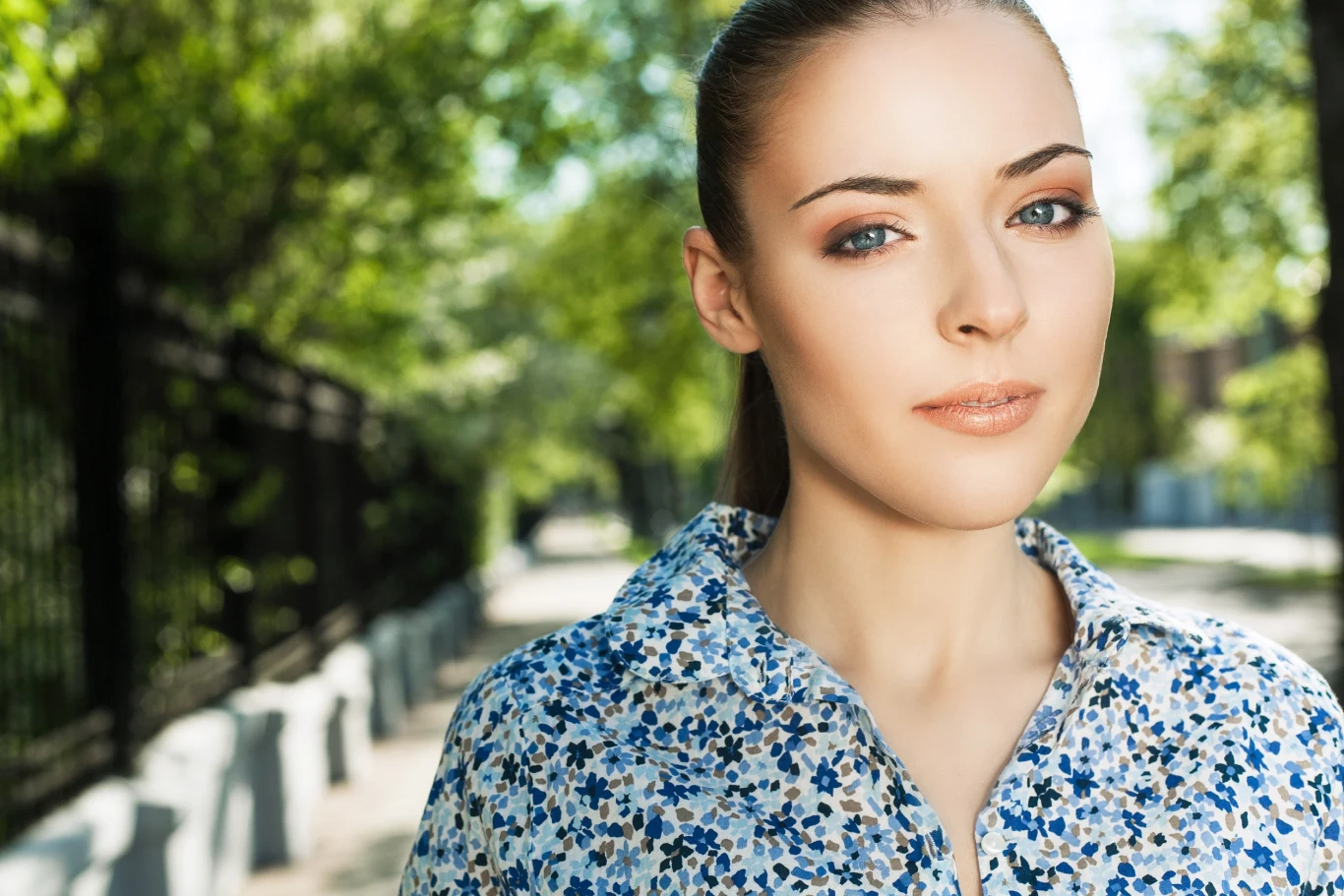 Młoda kobieta z jasną cerą i upiętymi włosami patrzy wprost, ubrana w koszulę w kwiatowy wzór, stojąca na tle zielonych drzew i ogrodzenia przy słonecznej, miejskiej alejce.