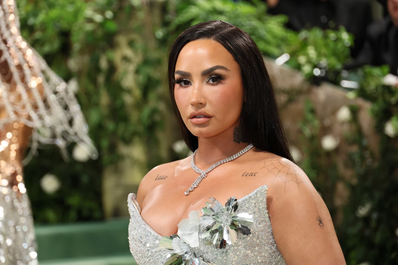 Demi Lovato Elegancko ubrana kobieta z długimi, ciemnymi włosami w błyszczącej, srebrnej sukni z kwiatową aplikacją, ozdobiona naszyjnikiem oraz subtelnym makijażem na tle dekoracji roślinnych.