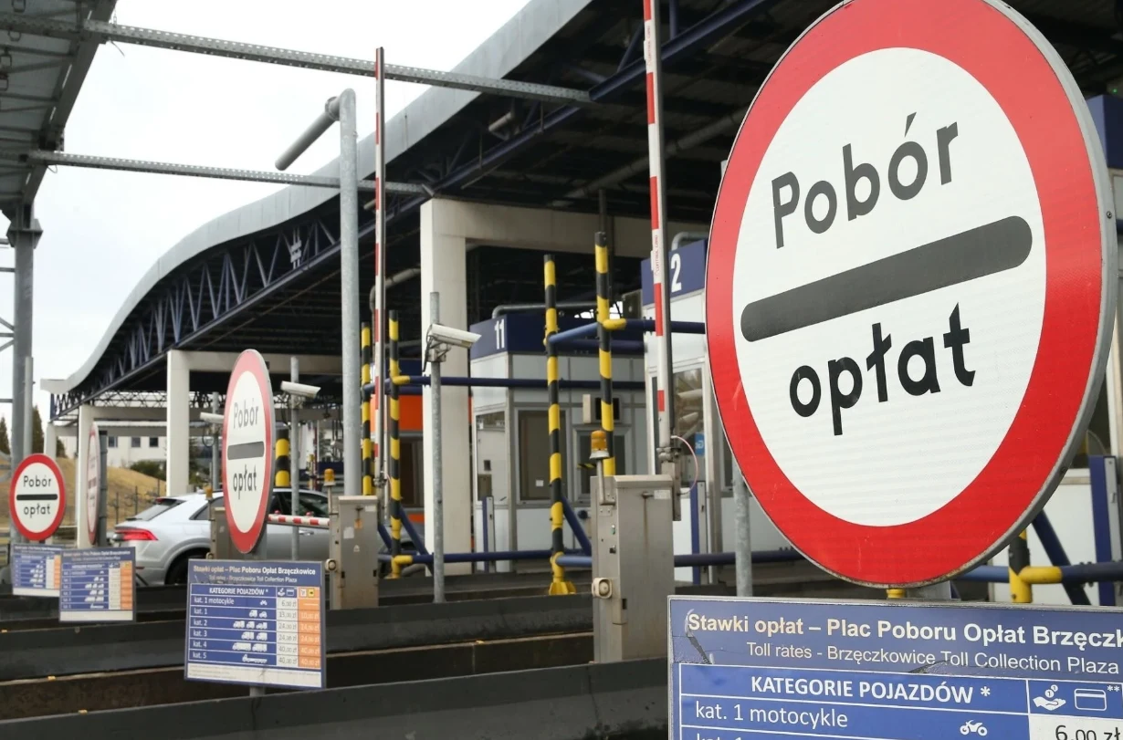 System e-TOLL przestał obowiązywać dla aut osobowych w połowie 2023 r., ale kary wciąż są naliczane. Plac poboru opłat z wyraźnymi znakami informującymi o konieczności uiszczenia opłaty oraz tablicami z cennikiem i kategoriami pojazdów, na drugim planie widoczne bramki i fragment samochodu.