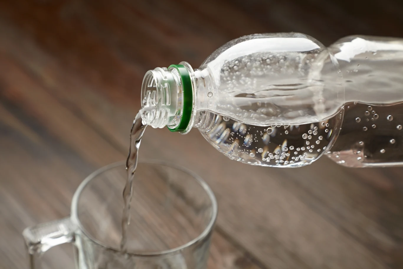 Krystynka - naturalna woda mineralna gazowana może zawierać kawałki szkła - ostrzega GIS / zdjęcie ilustracyjne Strumień wody wlewany z plastikowej butelki do przezroczystej szklanki, tło jest rozmyte i drewniane.