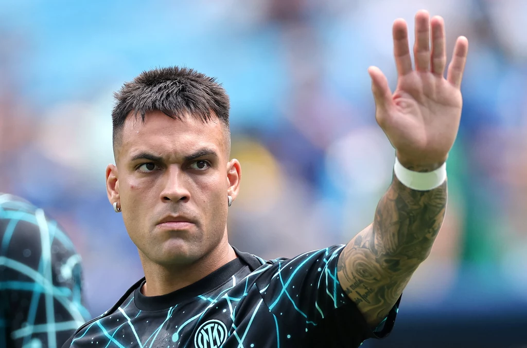 Lautaro Martinez Młody mężczyzna w sportowej koszulce z widocznym tatuażem na ramieniu unosi rękę w geście powitania lub pozdrowienia, w tle rozmyta, stadionowa sceneria.