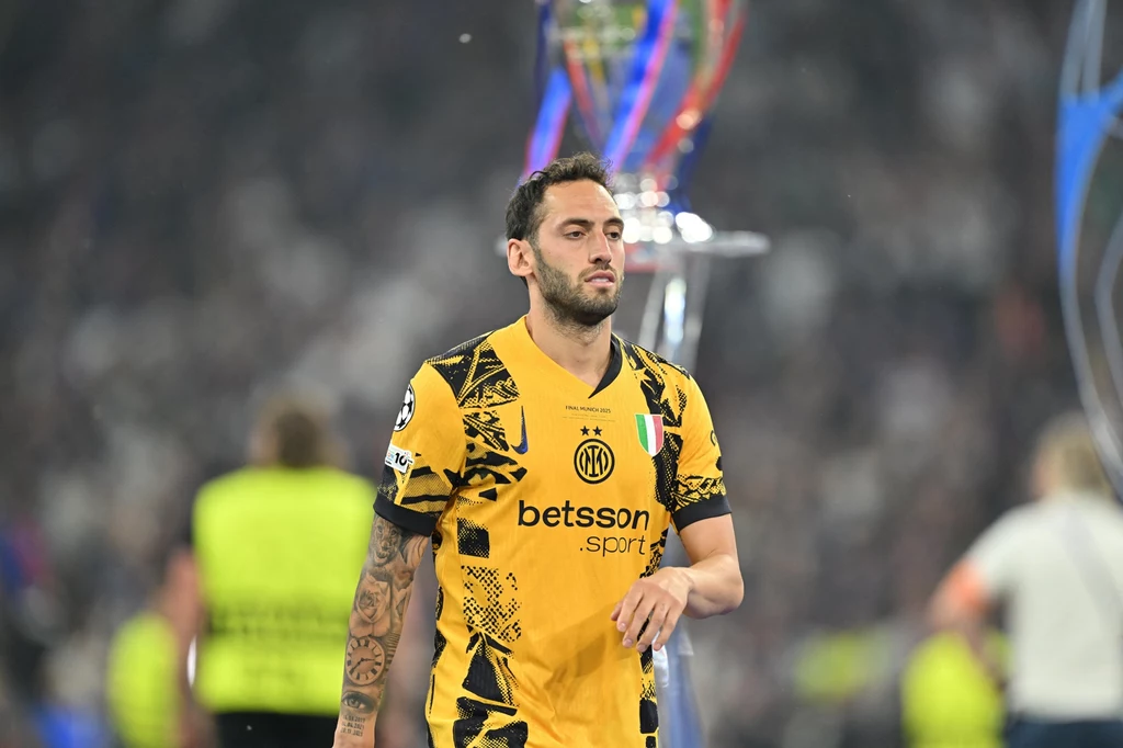 Hakan Calhanoglu Piłkarz w żółtej koszulce Interu Mediolan stoi na boisku, w tle duży puchar piłkarski oraz rozmyty tłum ludzi.