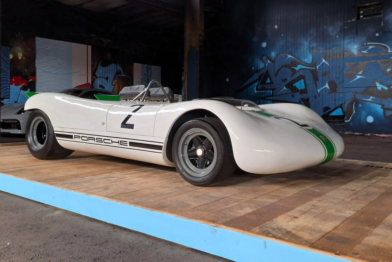 Klasyczny samochód wyścigowy Porsche o białym nadwoziu z zielonymi detalami i numerem 2 na drzwiach, stoi na drewnianym podeście w hali z niebieskim graffiti na ścianie w tle.