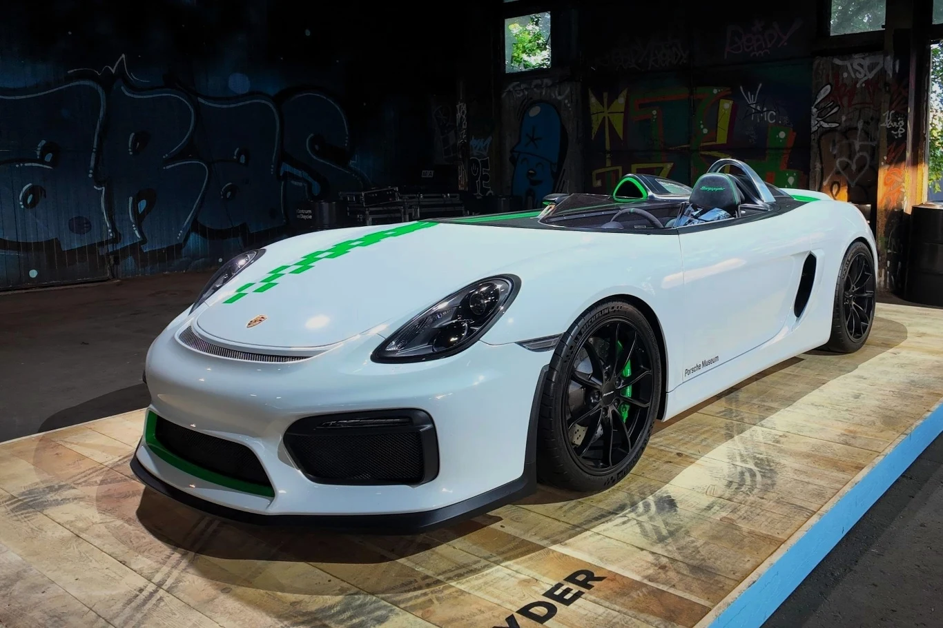 Biały sportowy samochód Porsche Boxster Spyder z zielonymi detalami, ustawiony na drewnianej platformie, na tle ściany z graffiti i wyraźnym światłem wpadającym przez okna.