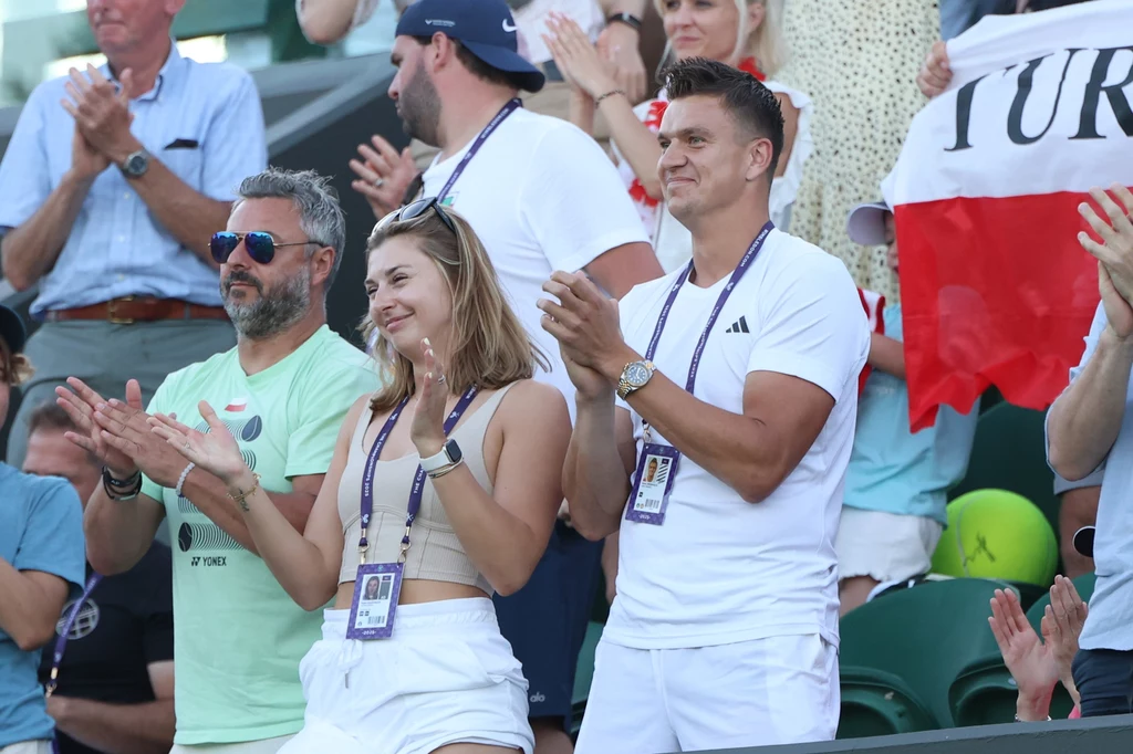 Bartek Witke, Marta Majchrzak i Karol Drzewiecki po wygraniu przez Kamila Majchrzaka meczu na Wimbledonie Grupa ludzi kibicująca na trybunach, w centrum kobieta i mężczyzna klaszczący z uśmiechami oraz druga osoba w okularach przeciwsłonecznych, w tle flaga Polski oraz tłum innych kibiców.