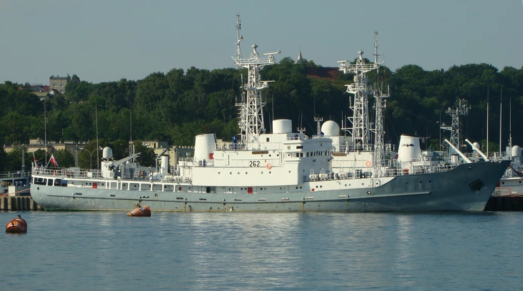 ORP Jerzy Różycki i ORP Henryk Zygalski mają zastąpić w polskiej marynarce okręty rozpoznania radioelektronicznego ORP Nawigator i ORP Hydrograf, które weszły do służby kolejno w 1975 i 1976 roku. Na zdjęciu ORP Nawigator Duży okręt cumujący przy nabrzeżu, wyposażony w liczne anteny i maszty, otoczony boiami na wodzie, w tle widoczne drzewa i budynki portowe.