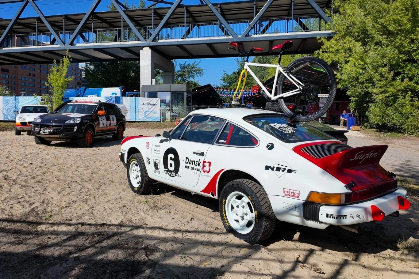 Samochód rajdowy Porsche 911 z numerem startowym 6 i rowerem zamocowanym na dachu stoi na piasku w pobliżu metalowej kładki dla pieszych, w tle dwa inne pojazdy rajdowe oraz zieleń drzew.