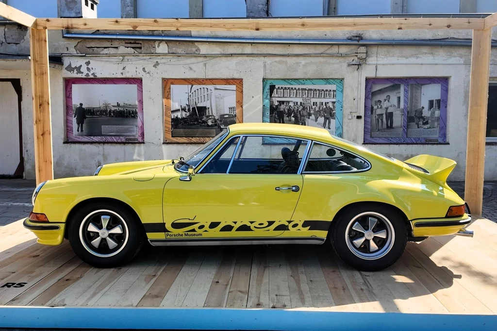 Porsche 911 Carrera RS 2.7 miało być zbudowane w liczbie tylko 500 egzemplarzy. Samochód cieszył się jednak takim zainteresowaniem, że niemiecki producent postanowił potroić produkcję. Żółty klasyczny samochód Porsche Carrera RS zaparkowany na drewnianym podeście, w tle ściana z czarno-białymi fotografiami w kolorowych ramach.