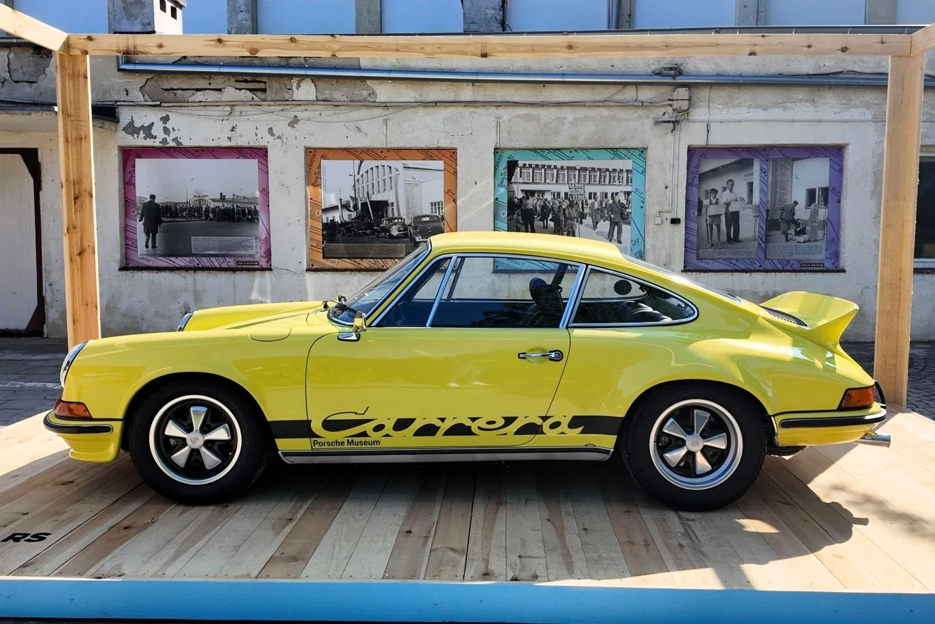 Żółty klasyczny samochód Porsche Carrera RS zaparkowany na drewnianym podeście, w tle ściana z czarno-białymi fotografiami w kolorowych ramach.