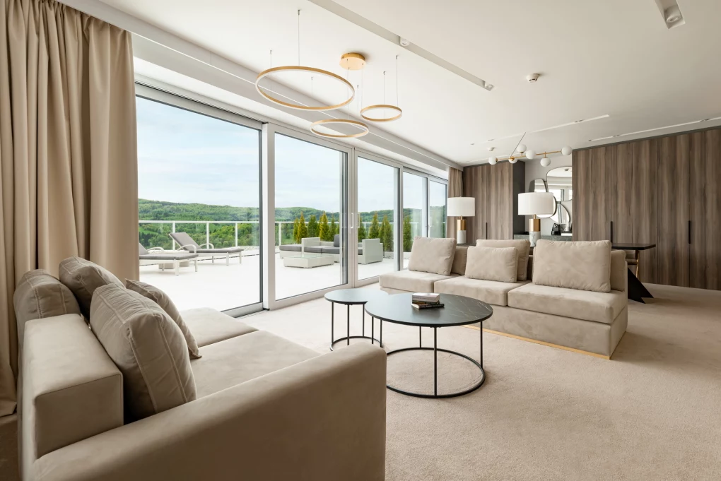 Hotel Crystal Mountain Przestronny salon z dużymi oknami sięgającymi od podłogi do sufitu, jasne beżowe sofy ustawione przy okrągłym stoliku kawowym, eleganckie lampy sufitowe oraz widok na taras z balustradą i zielonym krajobrazem za oknem.