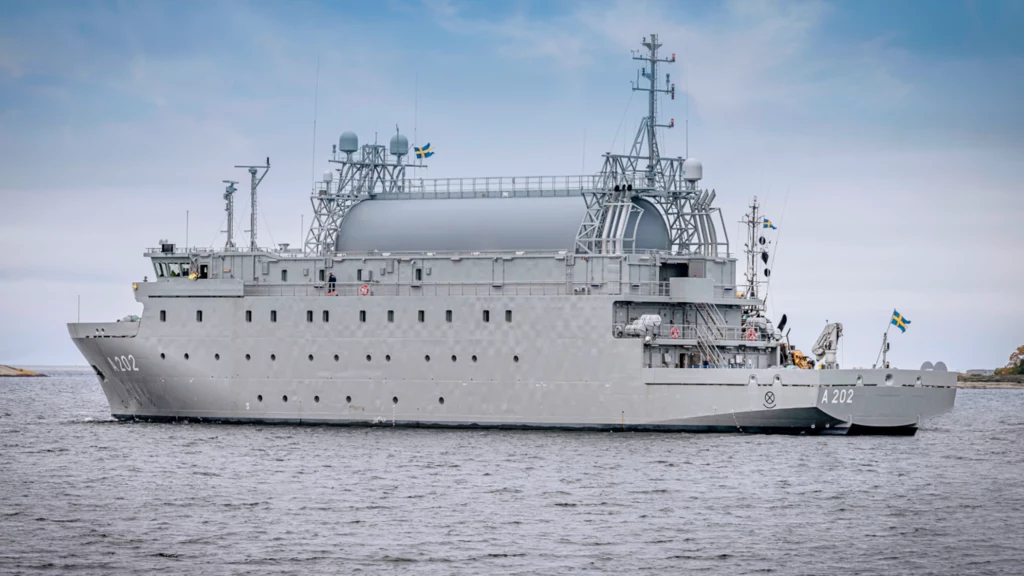 Okręt HMS Artemis Szwedzki okręt wojskowy w kolorze szarym płynie po spokojnych wodach, na pokładzie widoczne są maszty, anteny oraz szwedzka flaga na rufie; w tle fragment niewyraźnego wybrzeża i niebo z chmurami.