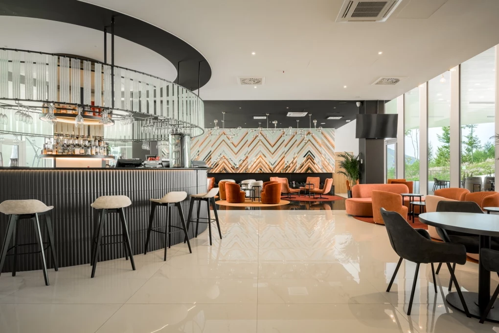 Hotel Crystal Mountain Nowoczesne wnętrze kawiarni z eleganckim barem otoczonym wysokimi stołkami, wygodne fotele w odcieniach pomarańczowego stoją przy okrągłych stolikach na tle przeszklonej ściany i geometrycznej dekoracji na dalszej ścianie.