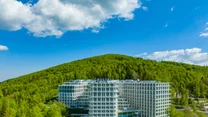 Nowoczesny, biały hotel Crystal położony na tle bujnego zielonego lasu i wzgórza, z basenem zewnętrznym oraz czystym niebem z białymi chmurami ponad budynkiem.