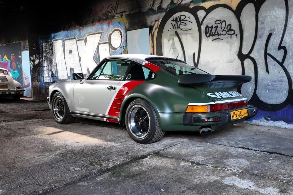 Porsche 930 Turbo stylizowane na samochód Johnny'ego Silverhanda z gry Cyberpunk 2077. Samochód sportowy Porsche 911 w dwukolorowym lakierze z naklejkami parkuje na tle kolorowych graffiti na ścianie, w opuszczonym, miejskim otoczeniu.