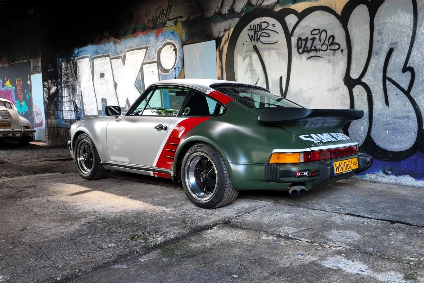 Samochód sportowy Porsche 911 w dwukolorowym lakierze z naklejkami parkuje na tle kolorowych graffiti na ścianie, w opuszczonym, miejskim otoczeniu.