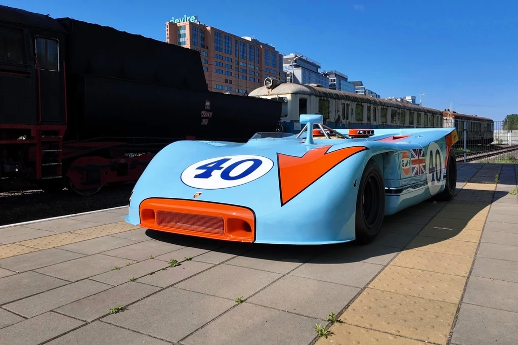 Między pociągami ukryte zostało Porsche 908/3 Spyder z 1970 roku. Ważący zaledwie 545 kg samochód generował 350 KM. Jasnoniebieski samochód wyścigowy z numerem 40 i pomarańczową strzałką stoi na peronie kolejowym, w tle znajdują się wagony pociągu oraz nowoczesny budynek