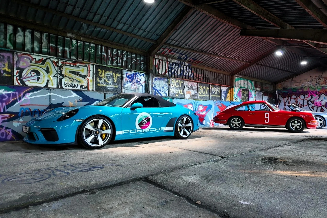Dwa sportowe samochody Porsche ustawione w opuszczonym garażu z kolorowymi graffiti na ścianach, nowoczesny model w kolorze niebieskim i klasyczny czerwony samochód z numerem 9