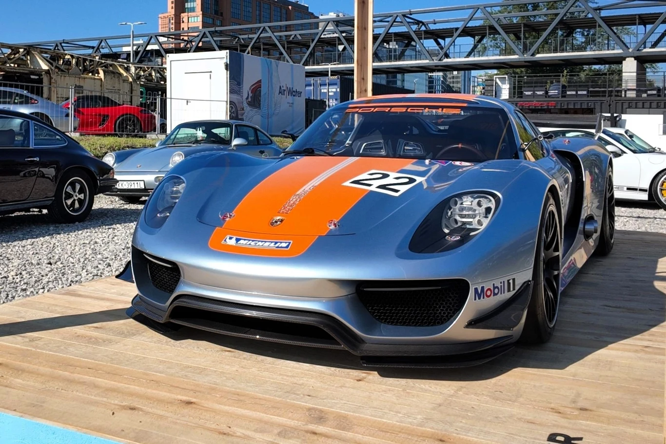 Sportowy samochód Porsche 918 Spyder z jasnopomarańczowym pasem i numerem 22 na masce stoi na drewnianym podeście, otoczony innymi samochodami oraz elementami infrastruktury miejskiej w tle.