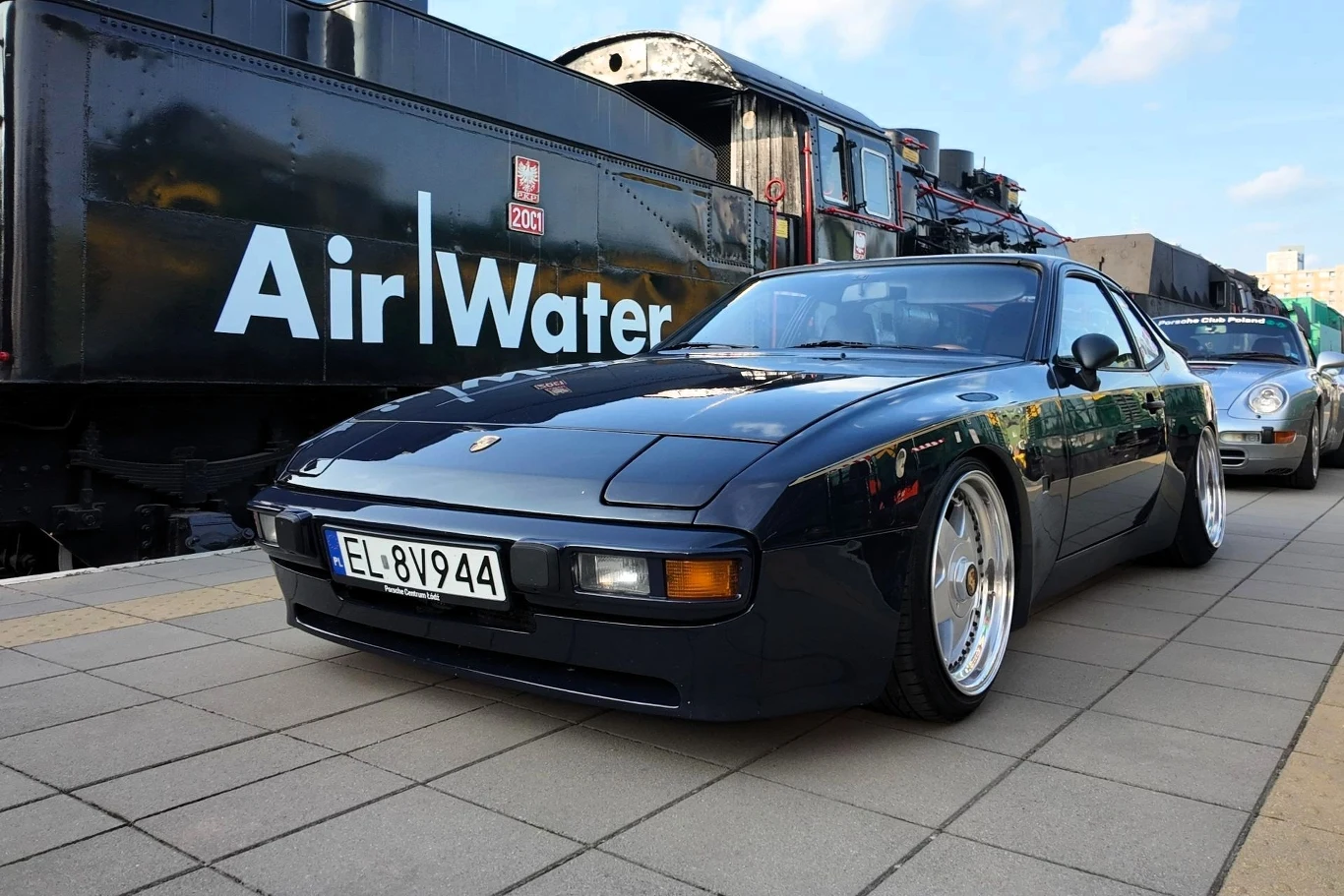 Czarny sportowy samochód marki Porsche zaparkowany na peronie obok zabytkowej lokomotywy z wyraźnym napisem „Air Water” na boku, w tle fragment innego pojazdu oraz fragment dworca kolejowego.