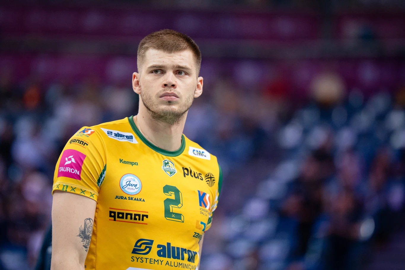 Młody siatkarz w żółtej koszulce klubowej z numerem 2 i licznymi logotypami sponsorów na tle rozmytej hali sportowej, skupiony wyraz twarzy, dynamiczna atmosfera meczu.