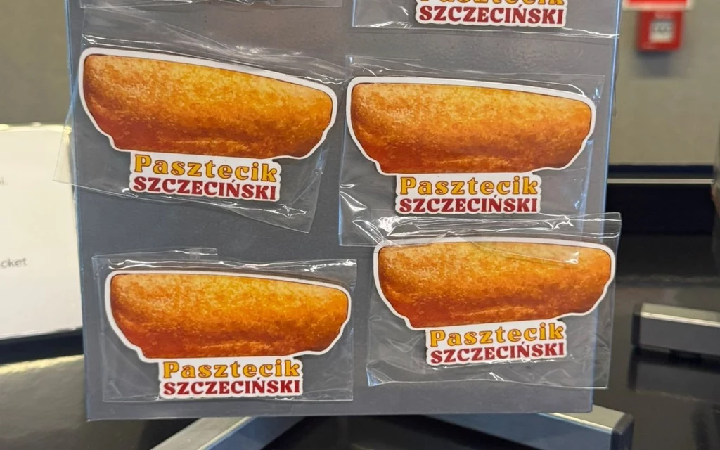 Naklejki przedstawiające pasztecika szczecińskiego, umieszczone na szarej planszy ekspozycyjnej, każda naklejka podpisana kolorowym napisem Pasztecik Szczeciński.
