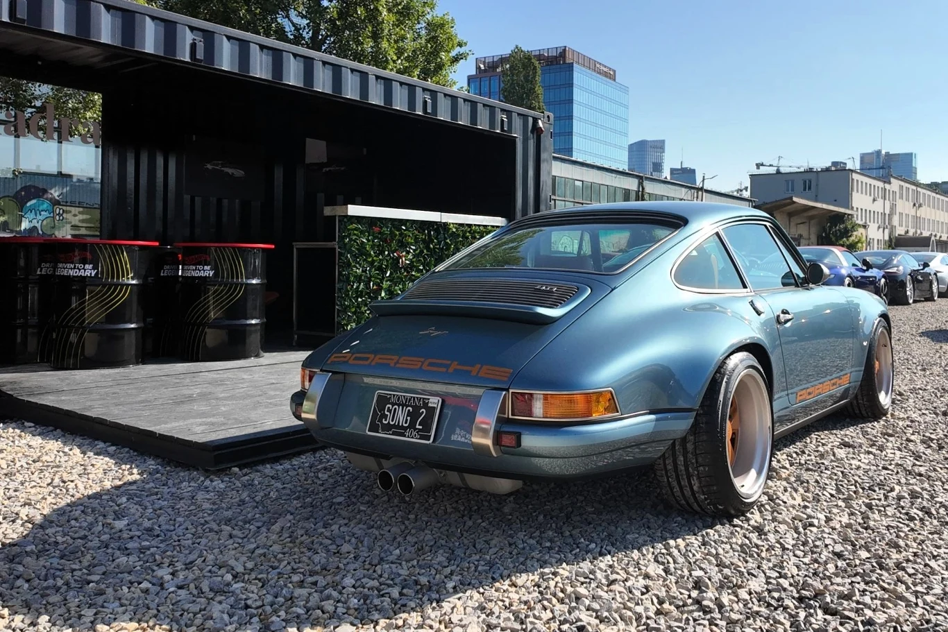 Klasyczny samochód Porsche o niebieskim kolorze stoi na żwirowym placu w pobliżu nowoczesnego kontenera, na tle miejskiej zabudowy i wieżowców.