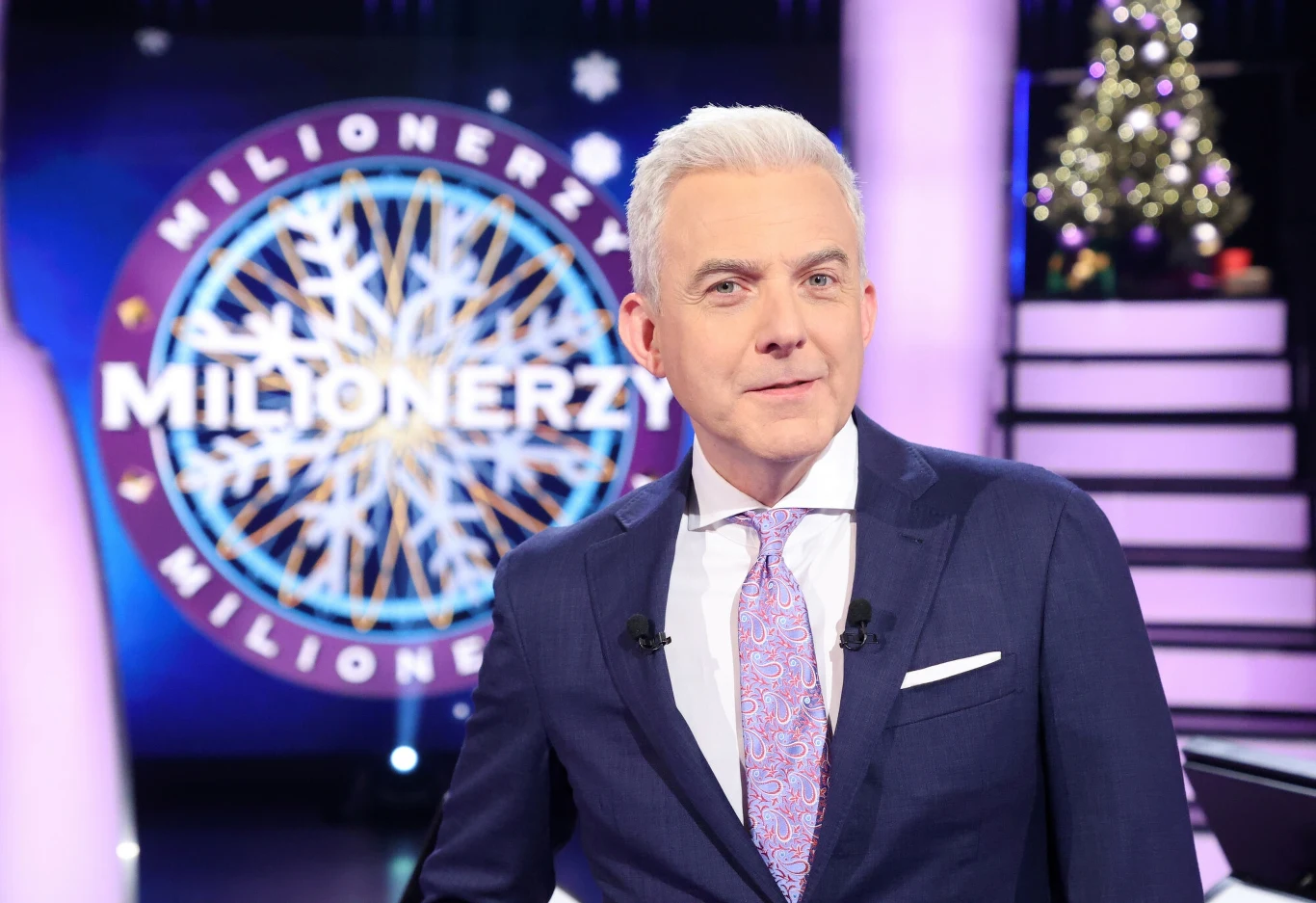 Mężczyzna w eleganckim garniturze i krawacie stoi w studio telewizyjnym przed charakterystycznym logo quizu Milionerzy, w tle widoczna jest dekoracja świąteczna.