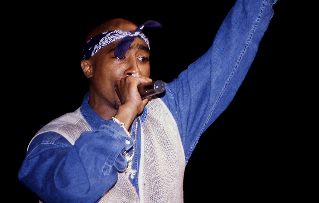Tupac Amaru Shakur Mężczyzna w niebieskiej bandanie na głowie i dżinsowej kurtce śpiewa do mikrofonu, unosząc jedną rękę do góry, wyraźnie zaangażowany w swój występ.