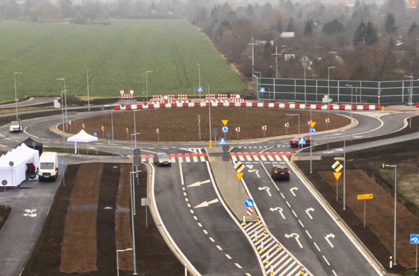 W poniedziałek marszałek woj. dolnośląskiego Paweł Gancarz podpisał z firmą Skanska umowę na budowę ostatniego odcinka wschodniej obwodnicy Wrocławia. nowoczesne rondo drogowe z wyraźnie oznaczonymi pasami ruchu, znakami drogowymi i barierami ochronnymi, kilka samochodów poruszających się po jezdni, w tle zieleń oraz pole, po lewej stronie biały namiot i samochód serwisowy