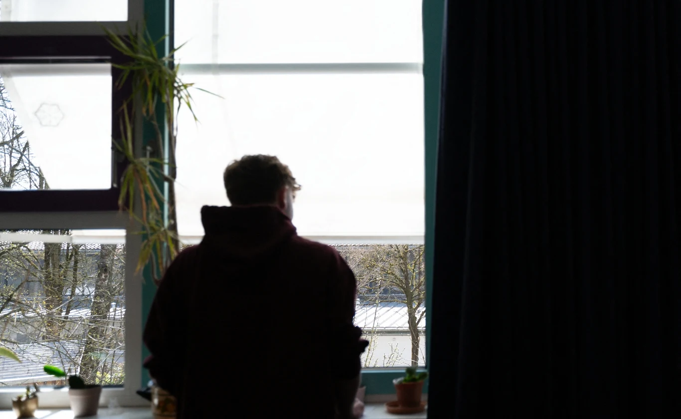 Niedzielne lęki. Dlaczego pod koniec weekendu odczuwamy niepokój? Sylwetka osoby stojącej tyłem przy oknie, patrzącej na zimowy krajobraz z drzewami, na parapecie kilka doniczkowych roślin, po lewej stronie widoczna wysoka roślina w doniczce.