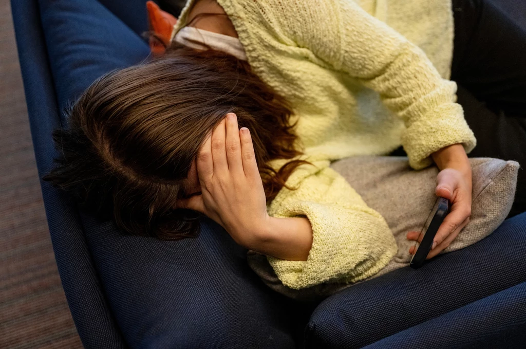 Kobieta leżąca na ciemnej kanapie z głową opartą o podłokietnik i dłonią zasłaniającą twarz, w drugiej ręce trzymająca telefon.