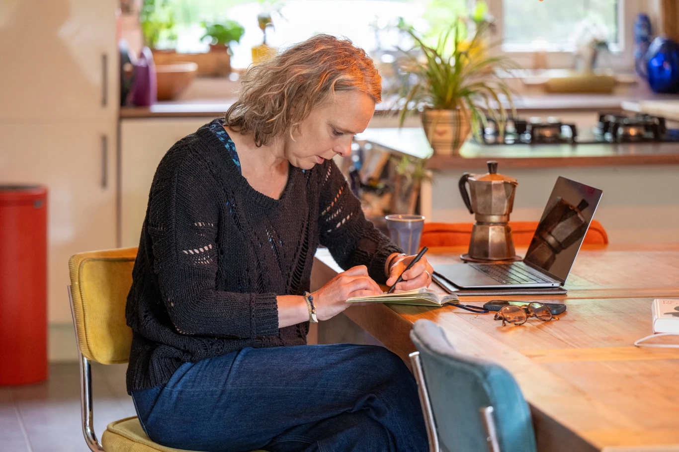 Przygotowanie "listy zadań" może pomóc zminimalizować niedzielny niepokój Kobieta z jasnymi falowanymi włosami siedzi przy stole w kuchni, pisze coś w notesie; obok stoi otwarty laptop, filiżanka i kawiarka, na stole leżą także okulary, w tle widoczne jasne wnętrze kuchni z roślinami przy oknie.
