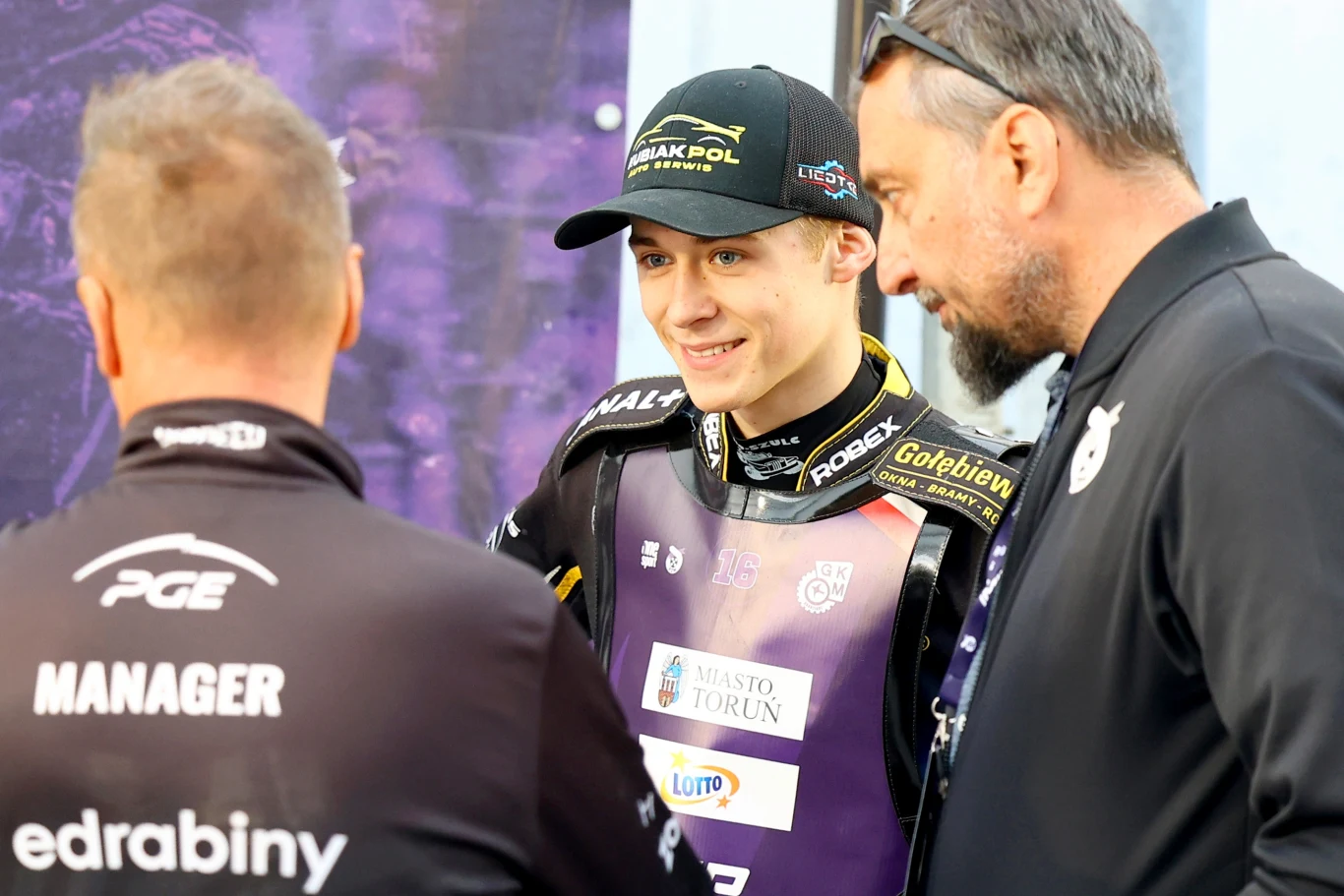 Młody mężczyzna w sportowym stroju i z czapką z daszkiem stoi uśmiechnięty w rozmowie z dwoma innymi mężczyznami, z których jeden ma na plecach napis 'MANAGER'.