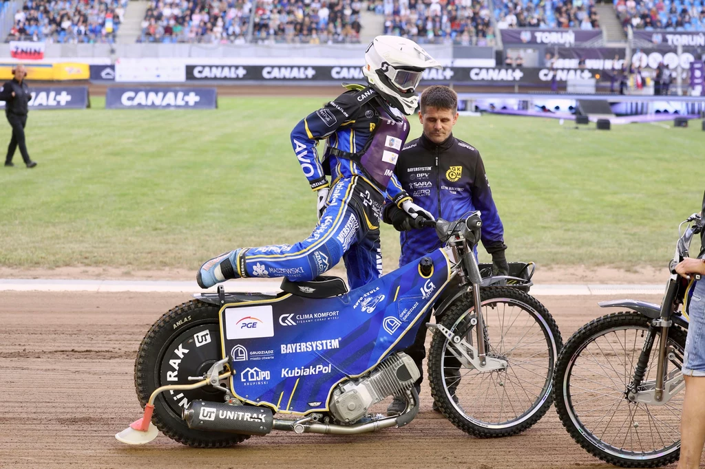 Kevin Małkiewicz szykuje się do startu. Żużlowiec w pełnym stroju ochronnym, przygotowujący się do startu, siedzi na motocyklu żużlowym na torze, obok stoi członek obsługi technicznej, a w tle widownia na stadionie.