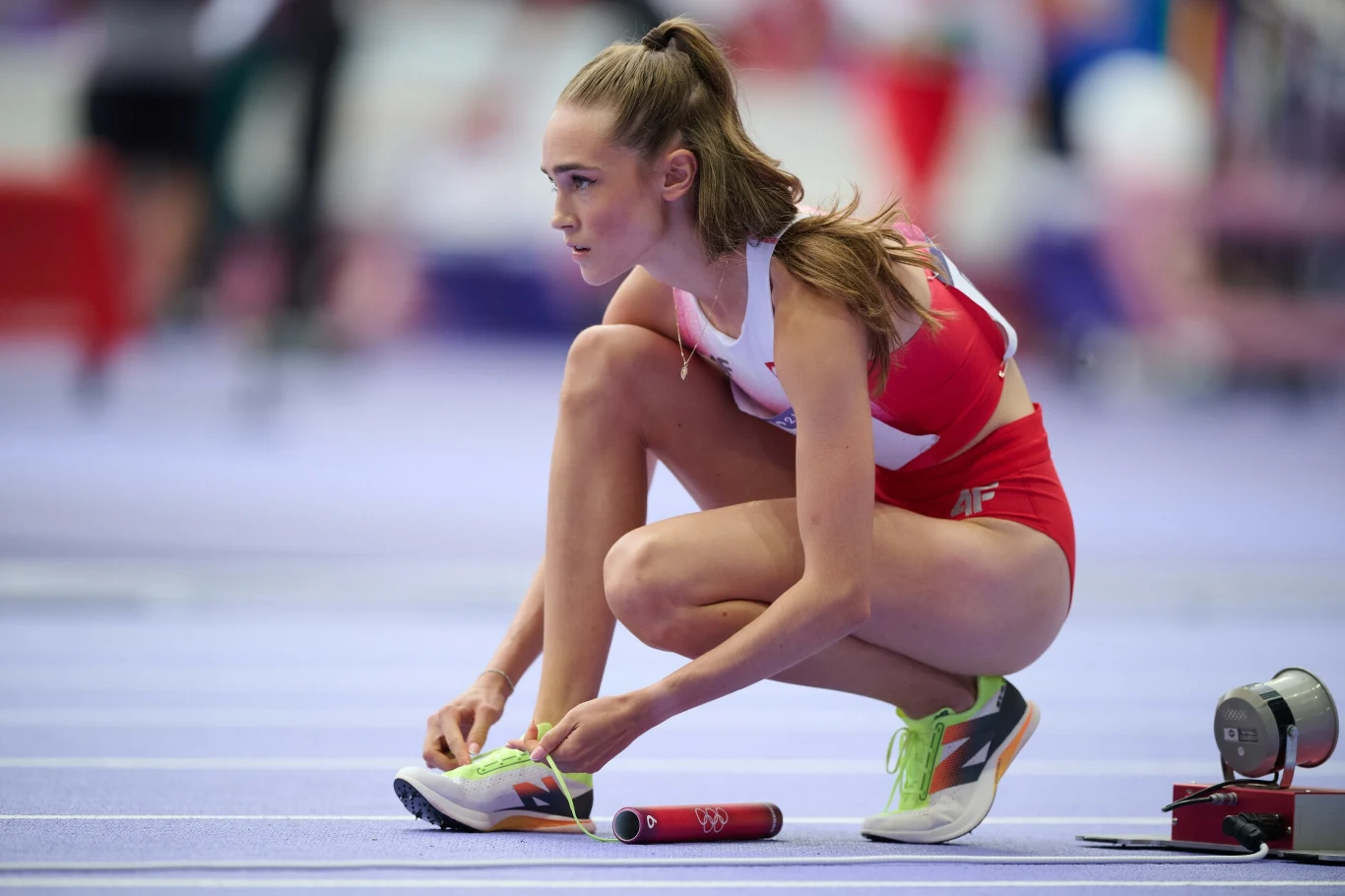 Lekkoatletka w czerwonym stroju sportowym wiąże but przed rozpoczęciem biegu sztafetowego na hali, trzymając pałeczkę w dłoni.