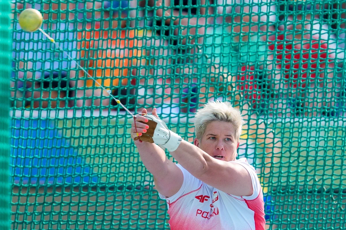 Lekkoatletka w biało-czerwonym stroju z napisem Polska wykonuje rzut młotem podczas zawodów, widoczna ruchoma piłka, zielona siatka ochronna w tle, skupiona twarz zawodniczki.