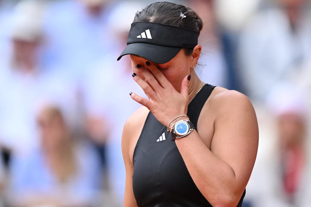 Zawodniczka tenisowa w sportowym stroju i czapce z daszkiem, z ręką na twarzy w geście emocji, na tle rozmytej publiczności.