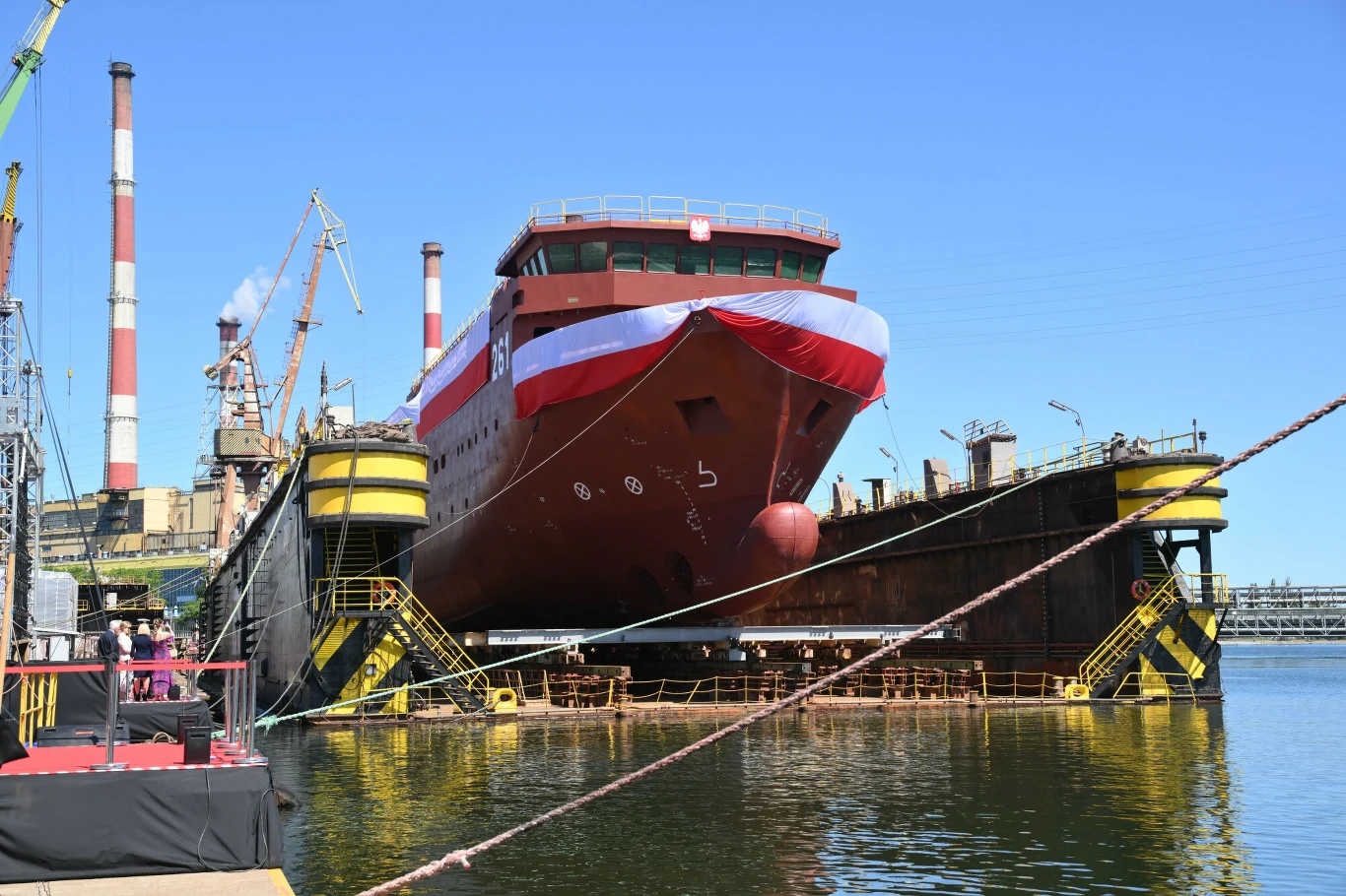 Coraz więcej chętnych do pracy w stoczni. Zatrudnienie ma wzrosnąć do 1200 osób Duży statek w stoczni podczas wodowania, ozdobiony biało-czerwoną flagą, stoi na pochylni otoczony infrastrukturą przemysłową, dźwigami oraz budynkami fabrycznymi w tle.