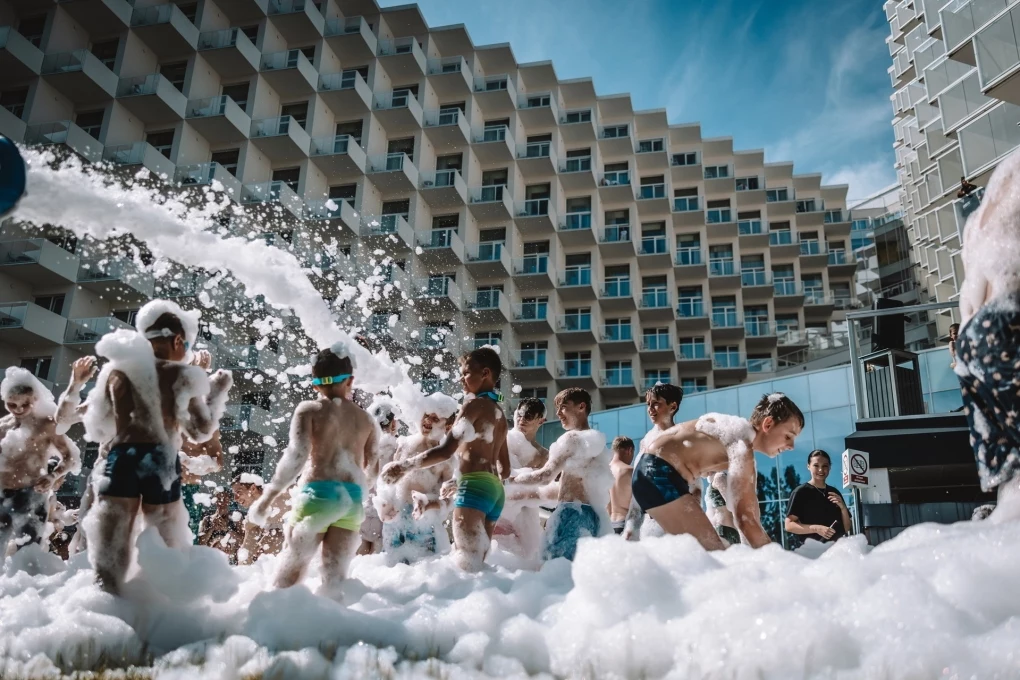 Hotel Crystal Mountain Dzieci bawiące się w pianie na tle nowoczesnego budynku, ubrane w stroje kąpielowe, z uśmiechami i radością podczas imprezy plenerowej w słoneczny dzień.
