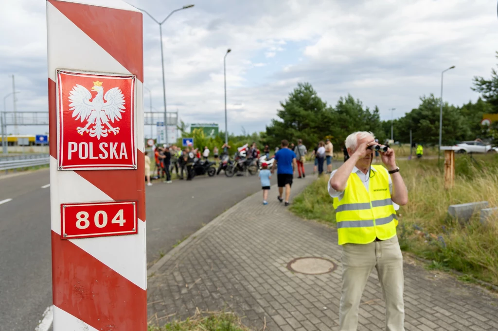 Słup graniczny Polski z godłem i napisem 'POLSKA' oraz numerem 804 stoi przy drodze asfaltowej. Na chodniku mężczyzna w żółtej kamizelce odblaskowej obserwuje okolicę przez lornetkę. W tle grupa osób, część z nich przy motocyklach, stoi na poboczu otoc...
