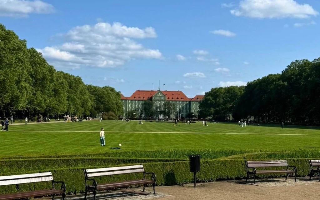 Duży, zadbany trawnik otoczony alejami drzew, w tle okazały budynek z czerwonym dachem, na środku ludzie spacerujący i bawiący się na świeżym powietrzu, ławki ustawione wzdłuż alejek.